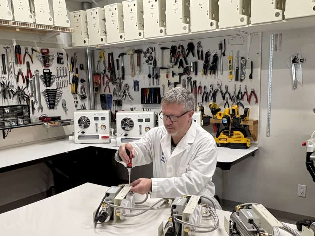 Technician in a lab coat repairs equipment at a workbench surrounded by tools and machinery in a workshop setting.